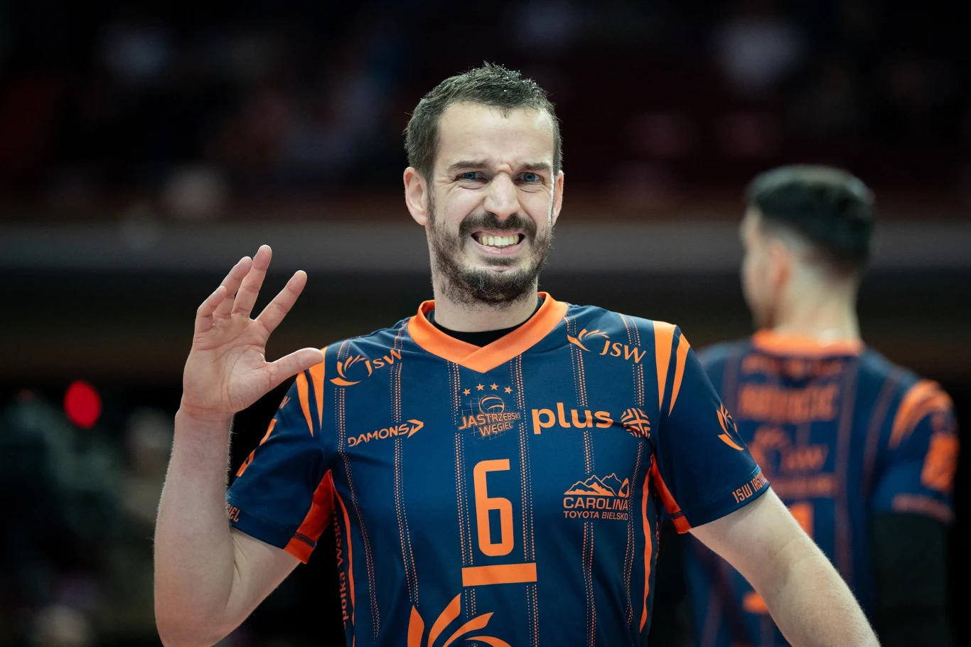 siatkarz w granatowo-pomarańczowej koszulce z numerem 6 wykonuje gest ręką i uśmiecha się podczas meczu, w tle widoczny drugi zawodnik