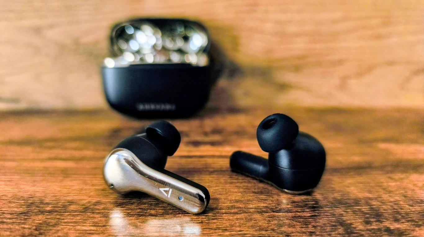 czarne bezprzewodowe słuchawki douszne z metalicznymi akcentami leżące na drewnianej powierzchni, w tle etui ładujące