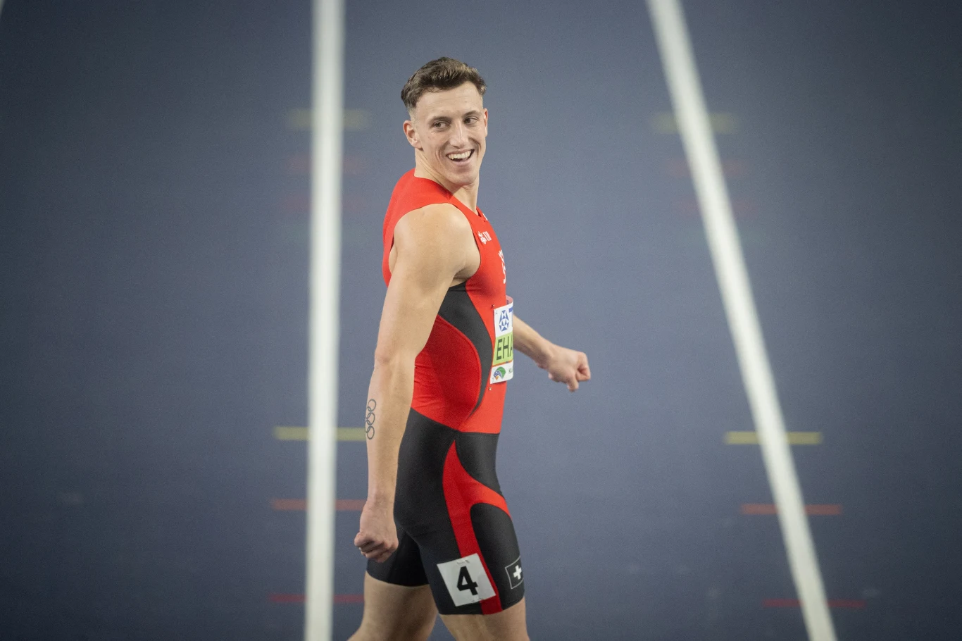 Lekkoatleta w czerwono-czarnym stroju startowym, noszący numer 4, odwraca się i uśmiecha na bieżni lekkoatletycznej.
