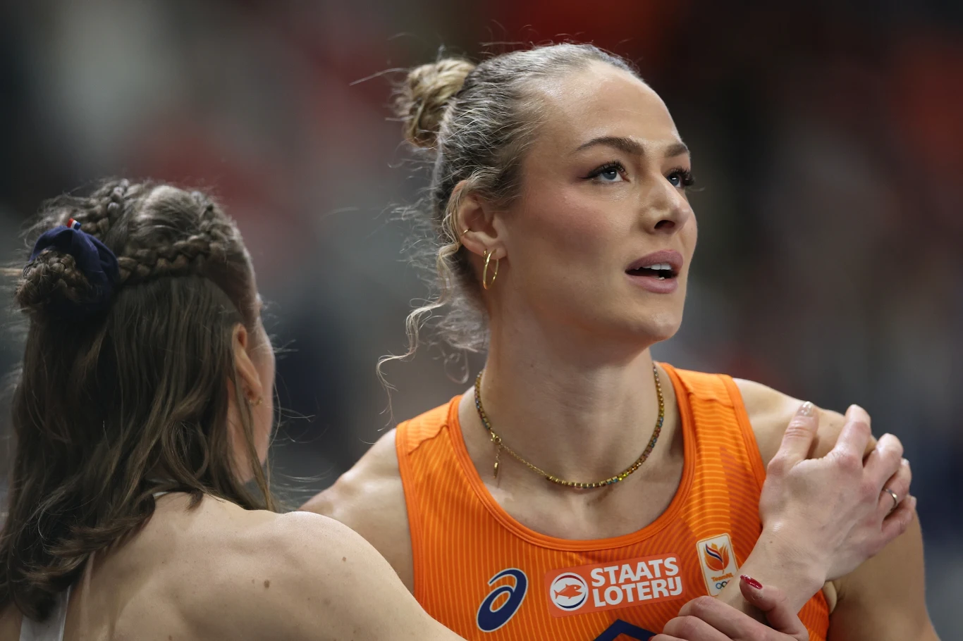 Lekkoatletka w pomarańczowym stroju z napisem 'STAATS LOTERIJ' odbiera gest wsparcia od drugiej kobiety po zakończonym biegu, wyraz skupienia i emocji na twarzy.