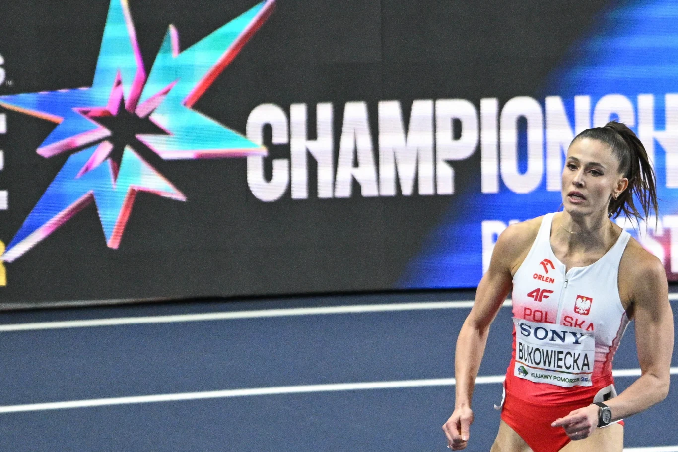 lekkoatletka w stroju reprezentacji Polski biegnie po bieżni podczas zawodów halowych, w tle widoczne logo mistrzostw na czarnym banerze