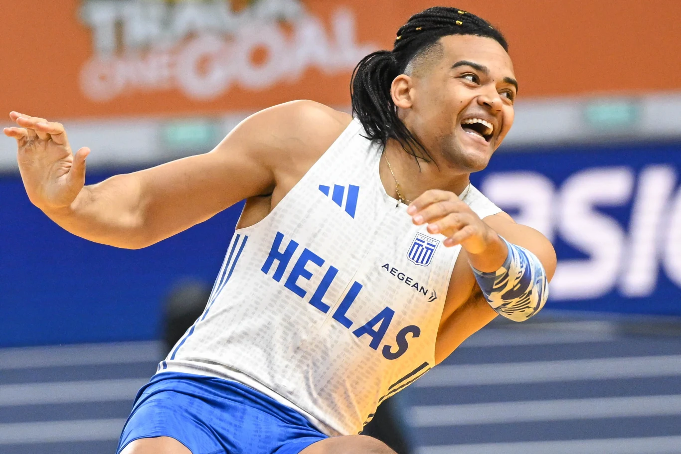 Emanuil Karalis sportowiec w białoniebieskim stroju z napisem Hellas wykonuje dynamiczny ruch podczas zawodów lekkoatletycznych, z wyrazem radości na twarzy i charakterystyczną fryzurą