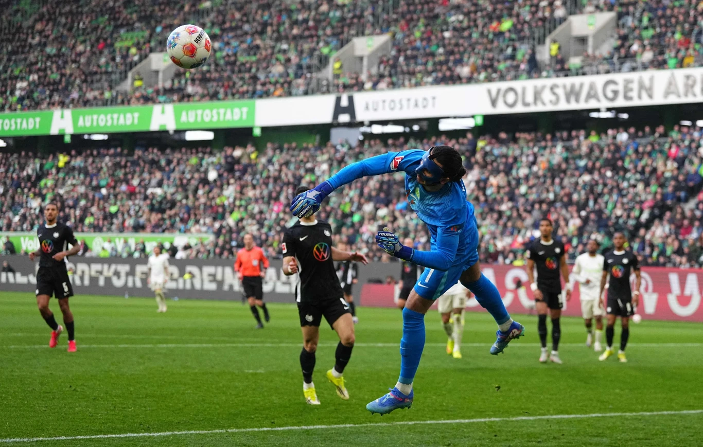 bramkarz w niebieskim stroju wykonuje wybicie piłki głową w polu karnym podczas meczu piłki nożnej, w tle piłkarze obu drużyn oraz pełne trybuny kibiców