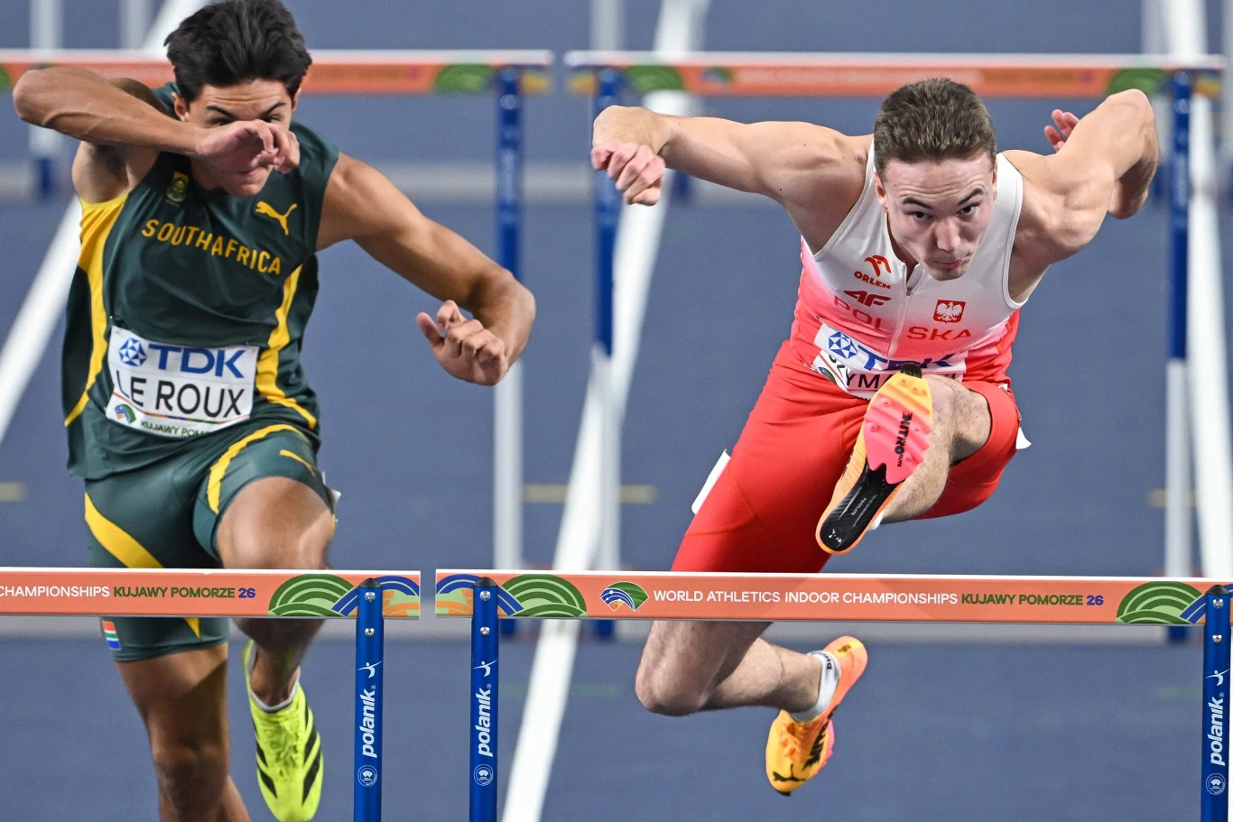 Dwóch lekkoatletów w trakcie biegu przez płotki intensywnie rywalizuje, przeskakując przeszkody; jeden ubrany w zielony strój z napisem South Africa, drugi w biało-czerwonym stroju reprezentacji Polski, mocno skoncentrowani na osiągnięciu mety.