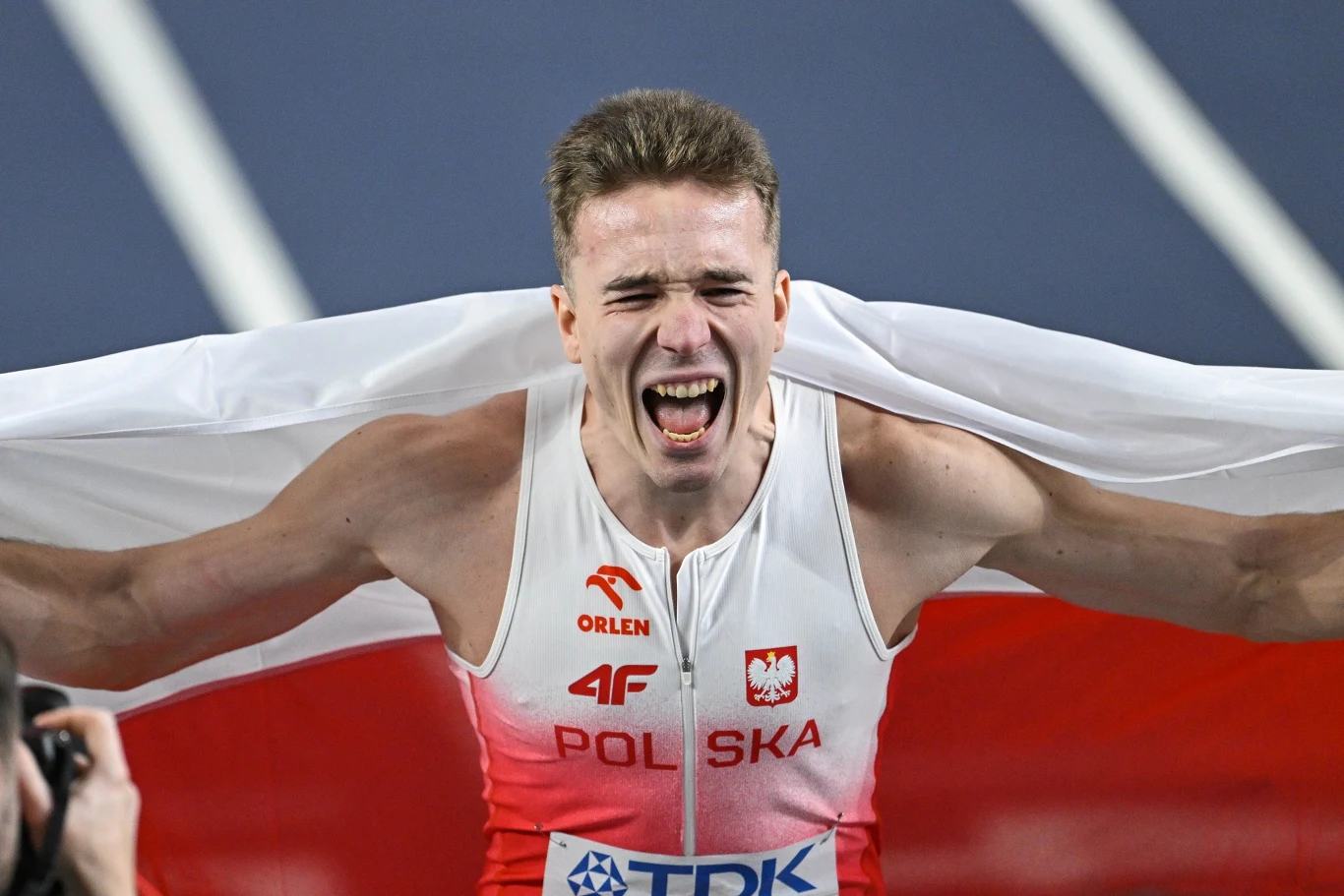 lekkoatleta w stroju z napisem 'Polska' i biało-czerwoną flagą na plecach wyraża emocje po udanym starcie na bieżni