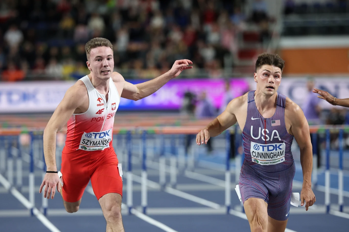Dwóch lekkoatletów finiszujących na bieżni podczas biegu płotkarskiego – po lewej zawodnik z Polski w biało-czerwonym stroju, po prawej zawodnik z USA w fioletowym stroju, obaj wyciągają ręce do przodu walcząc o zwycięstwo.