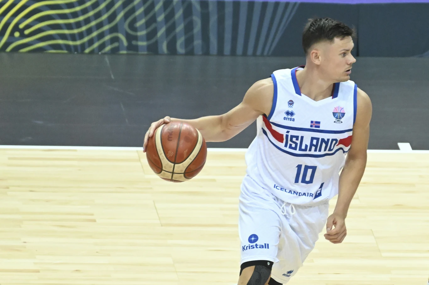 koszykarz w białym stroju drużyny Islandii z numerem 10 prowadzi piłkę do koszykówki po hali sportowej, skupiony na grze