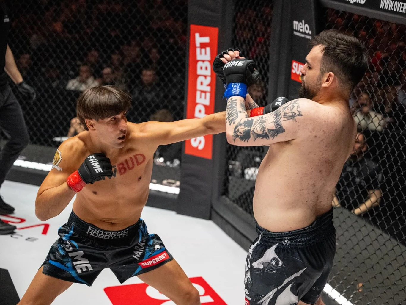 dwóch mężczyzn walczy w klatce MMA; jeden z nich uderza drugiego prostym lewym ciosem, podczas gdy przeciwnik z tatuażami na ramieniu broni się uniesioną ręką, a wokół klatki widać reklamy i ogrodzenie