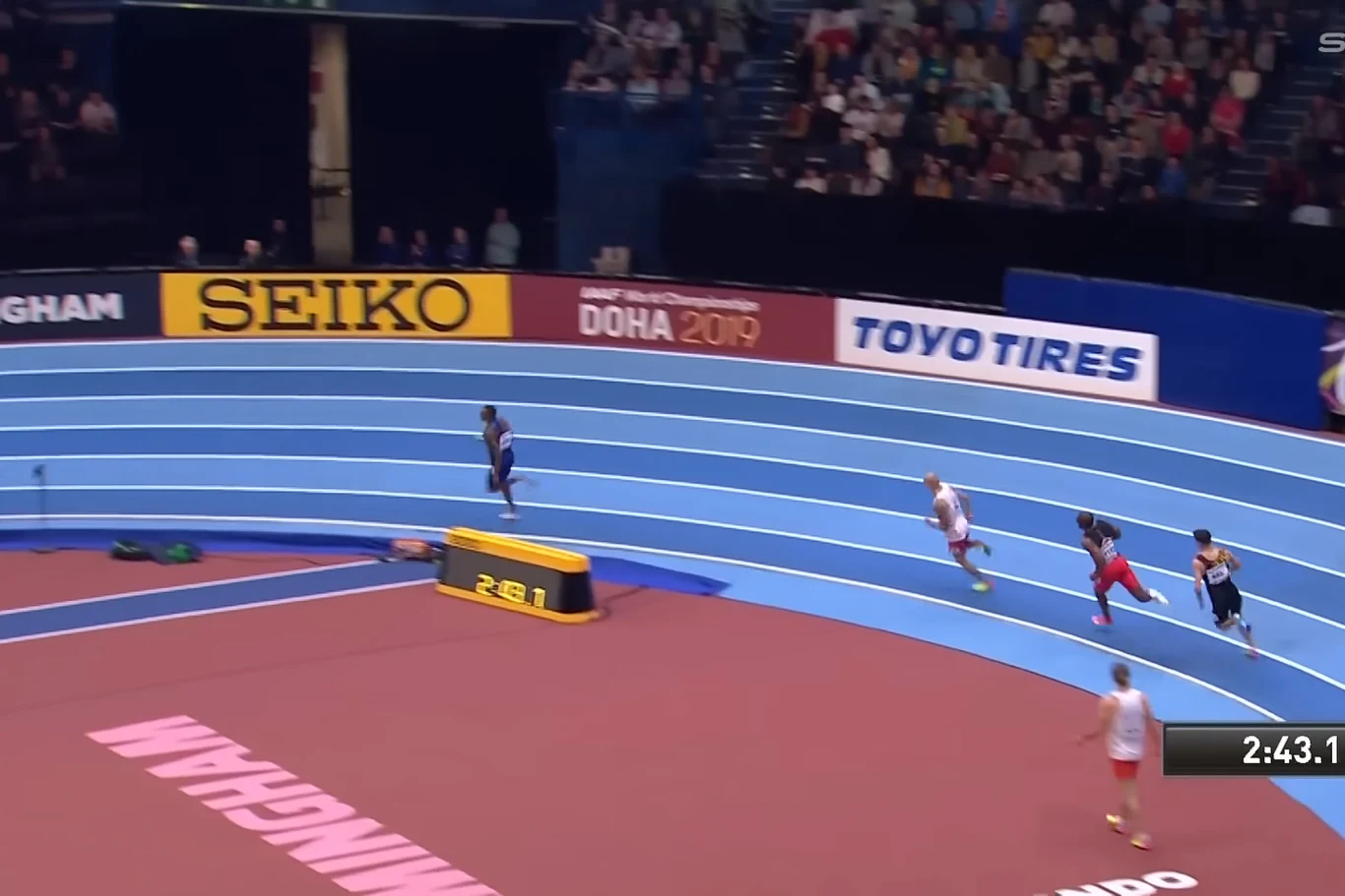 trzech lekkoatletów biegnie po niebieskiej bieżni na hali sportowej, jeden zawodnik wyraźnie prowadzi, a na trybunach znajduje się liczna widownia