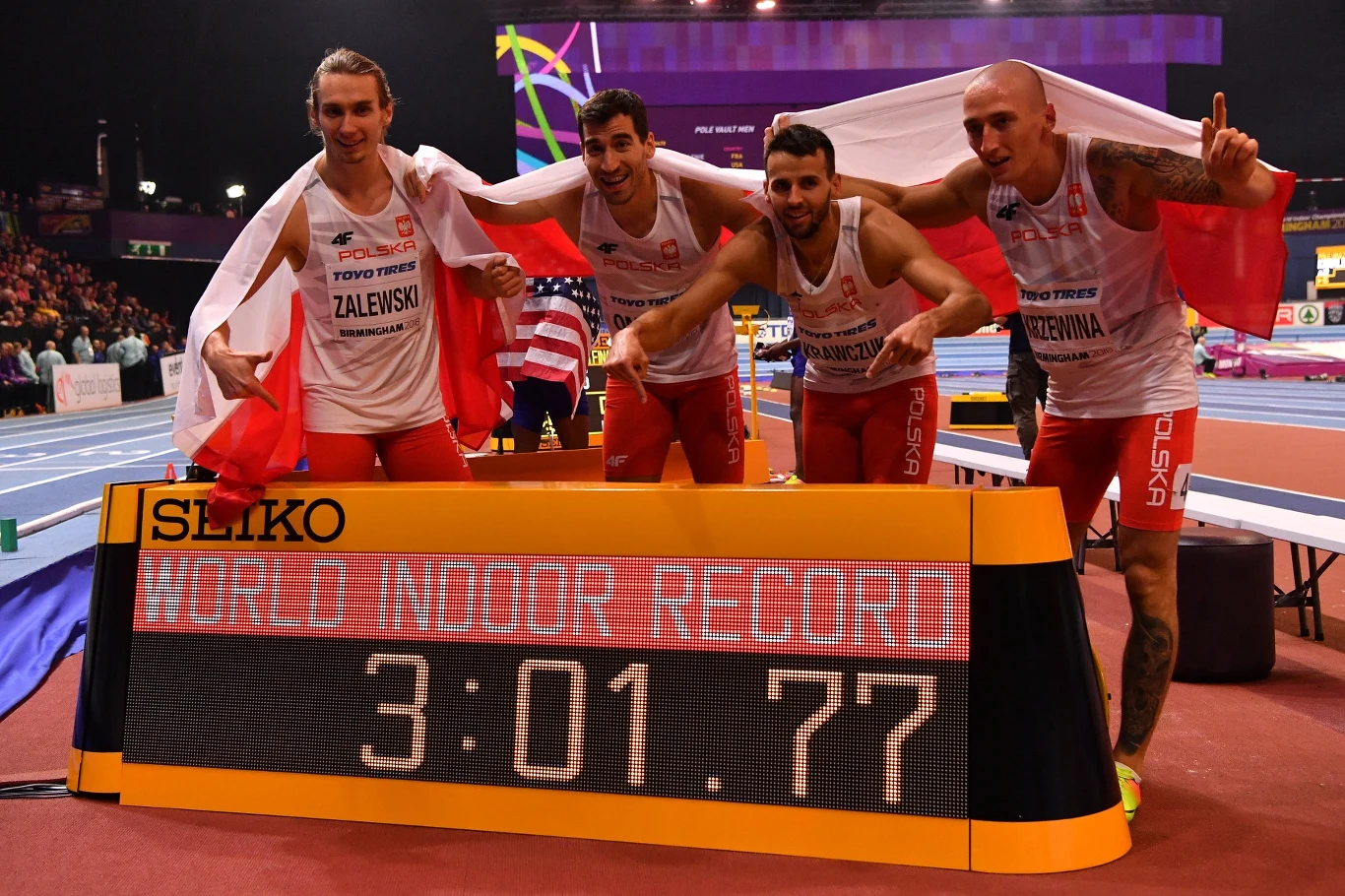 Czterech polskich lekkoatletów ubranych w stroje reprezentacji narodowej pozuje z flagą Polski obok elektronicznej tablicy z napisem 'WORLD INDOOR RECORD' i wynikiem 3:01.77, wyraźnie świętując ustanowienie rekordu świata w biegu sztafetowym.