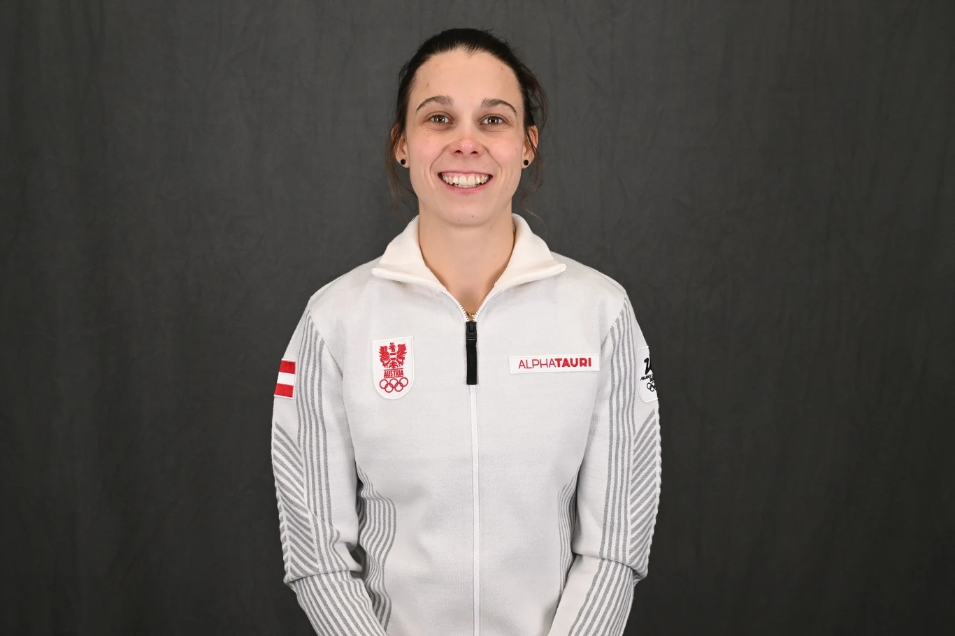 Kobieta w białej sportowej bluzie z logo olimpijskim oraz napisem Alphatauri, na tle ciemnej, jednolitej ściany, uśmiechnięta, z ciemnymi włosami spiętymi do tyłu.