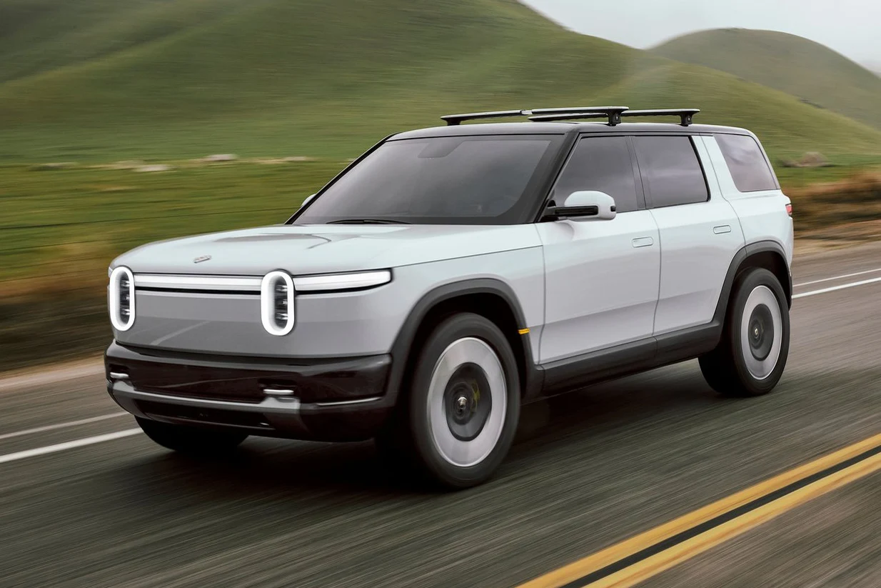 jasnoszary elektryczny SUV z charakterystycznymi okrągłymi światłami LED z przodu jedzie po asfaltowej drodze na tle zielonych pagórków