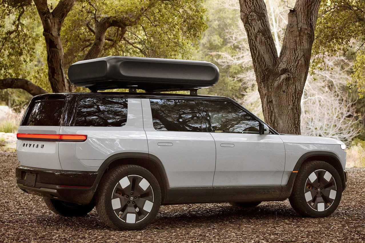 srebrny SUV z czarnym boksem dachowym zaparkowany pod dużymi drzewami w leśnym otoczeniu