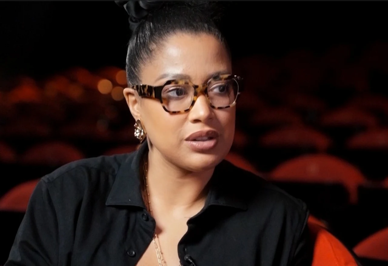 Patricia Kazadi Kobieta z ciemnymi upiętymi włosami i okularami w ciemnych oprawkach siedzi na tle rozmytych czerwonych siedzeń, ubrana w czarną koszulę i ozdobne kolczyki.