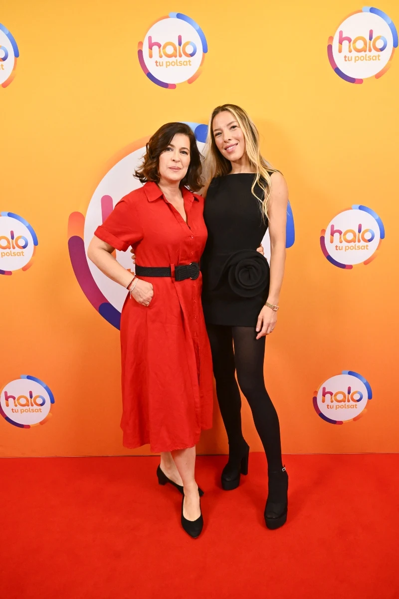 Dr hab. n. med. Anna Jeznach-Steinhagen i Ewa Chodakowska w "halo tu polsat" Dwie kobiety pozują razem na czerwonym dywanie przed pomarańczową ścianką z logotypami, jedna ubrana w czerwoną sukienkę z czarnym paskiem, druga w krótką czarną sukienkę z rajstopami i czarnymi butami na obcasie.