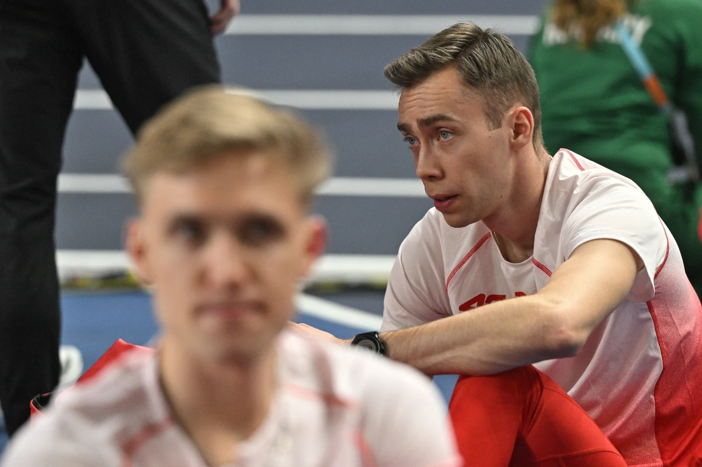 Dwóch młodych sportowców w biało-czerwonych strojach siedzi na bieżni, jeden z nich w ostrości wygląda na skupionego i zamyślonego, drugi rozmyty na pierwszym planie.