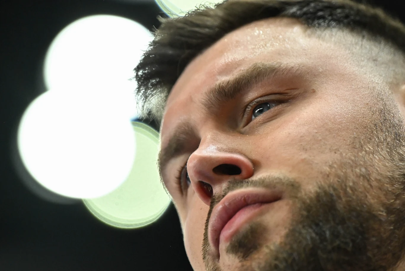 Zbliżenie na twarz młodego mężczyzny z zarostem i krótkimi włosami, z widocznymi drobnymi kroplami potu na czole, w tle rozmyte jasne światła.