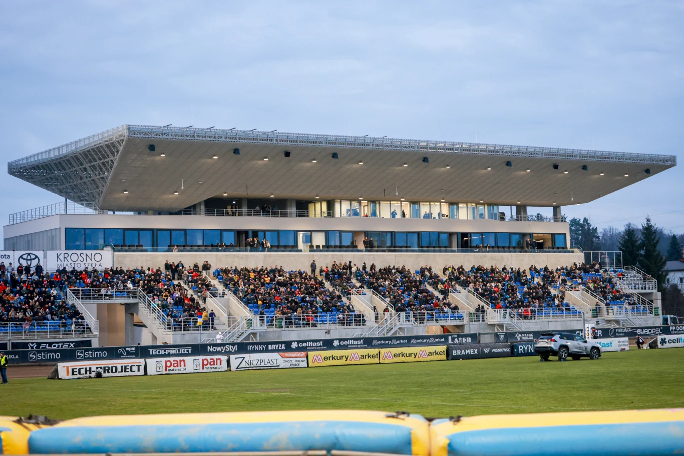 nowoczesna, wielopoziomowa trybuna stadionowa z zadaszeniem wypełniona publicznością, na pierwszym planie zielona murawa oraz samochód terenowy poruszający się wzdłuż trybun