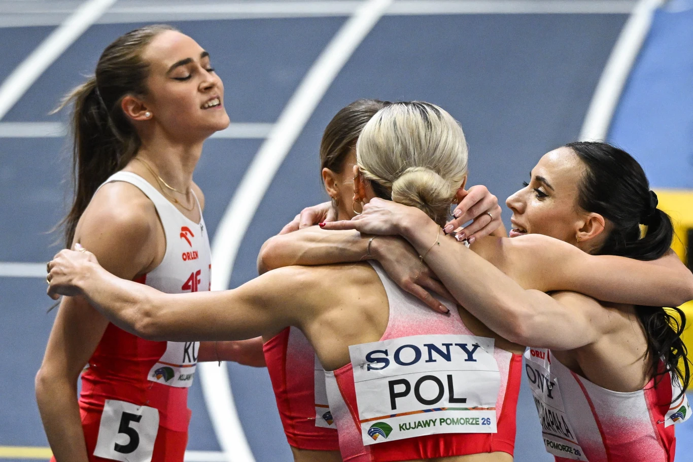 Cztery polskie lekkoatletki w biało-czerwonych strojach obejmują się na bieżni po ukończonym biegu, okazując radość i wzajemne wsparcie; jedna z zawodniczek ma numer 5, a na plecach drugiej widnieje napis 'POL'.