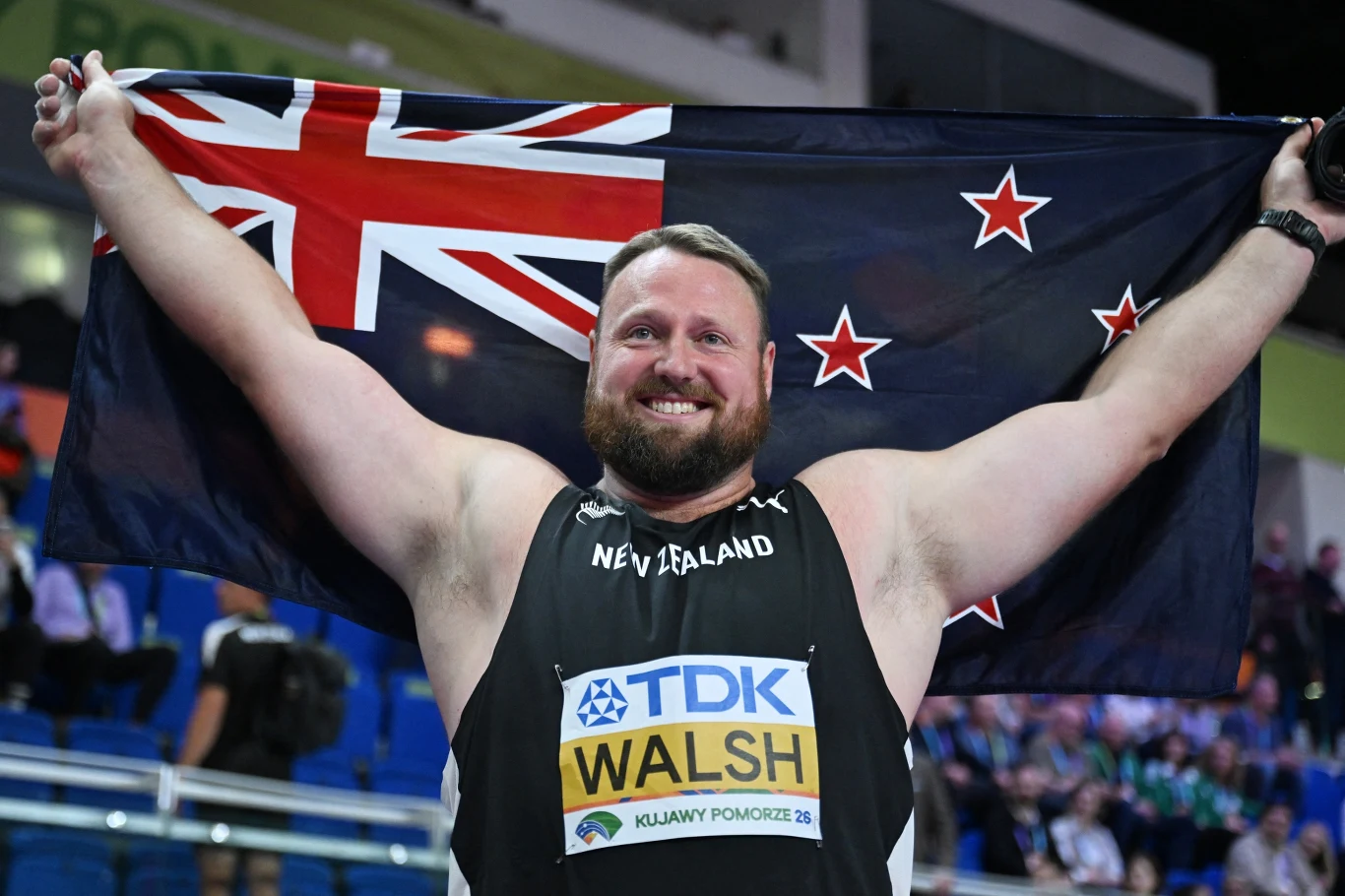Mężczyzna w sportowym stroju z napisem 'New Zealand' trzyma uniesioną flagę Nowej Zelandii, szeroko uśmiechnięty, stojąc na tle trybun pełnych kibiców.