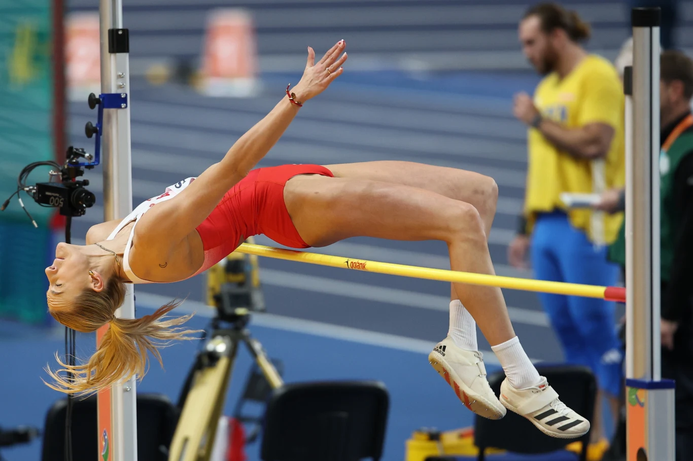 Lekkoatletka w trakcie skoku wzwyż unosi ciało ponad poprzeczkę, dynamiczna sylwetka w locie, sportowa hala w tle, skupienie na precyzji ruchu.