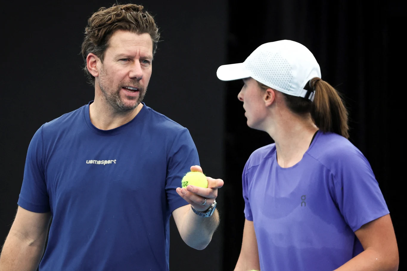 Trener omawia strategię z tenisistką, która trzyma rakietę tenisową i żółtą piłkę, oboje skoncentrowani na rozmowie podczas treningu na korcie.