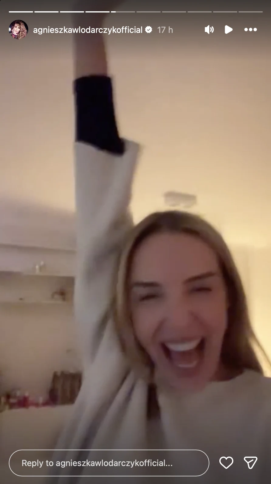 kobieta o długich, jasnych włosach z szerokim uśmiechem na twarzy unosi jedną rękę w górę, wyrażając radość i entuzjazm, w tle rozmyte, jasne wnętrze mieszkania