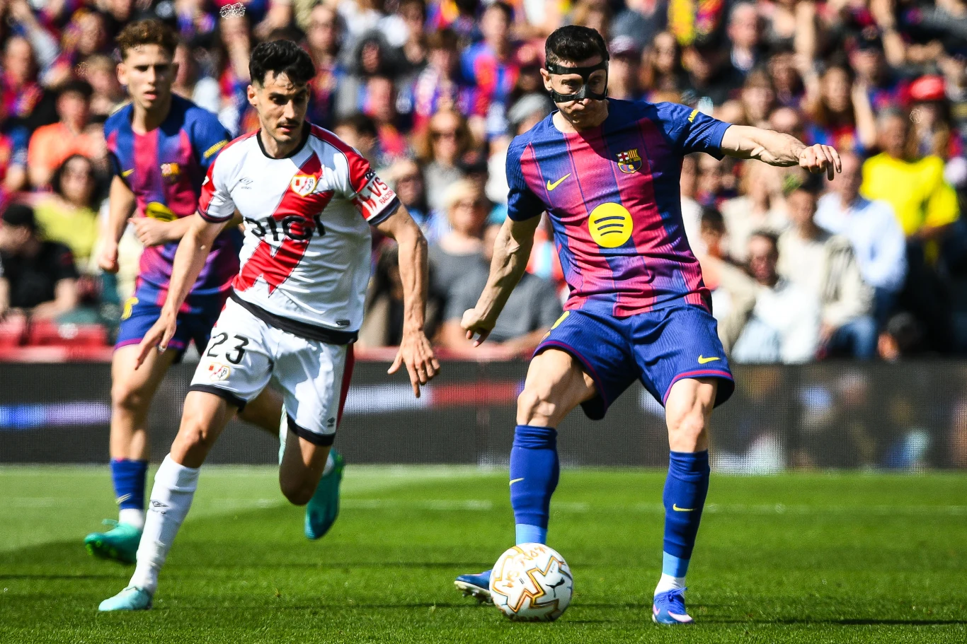 piłkarz w niebiesko-granatowej koszulce FC Barcelony kontroluje piłkę, ścigany przez zawodnika w biało-czerwonym stroju zespołu przeciwnego na tle trybun pełnych kibiców