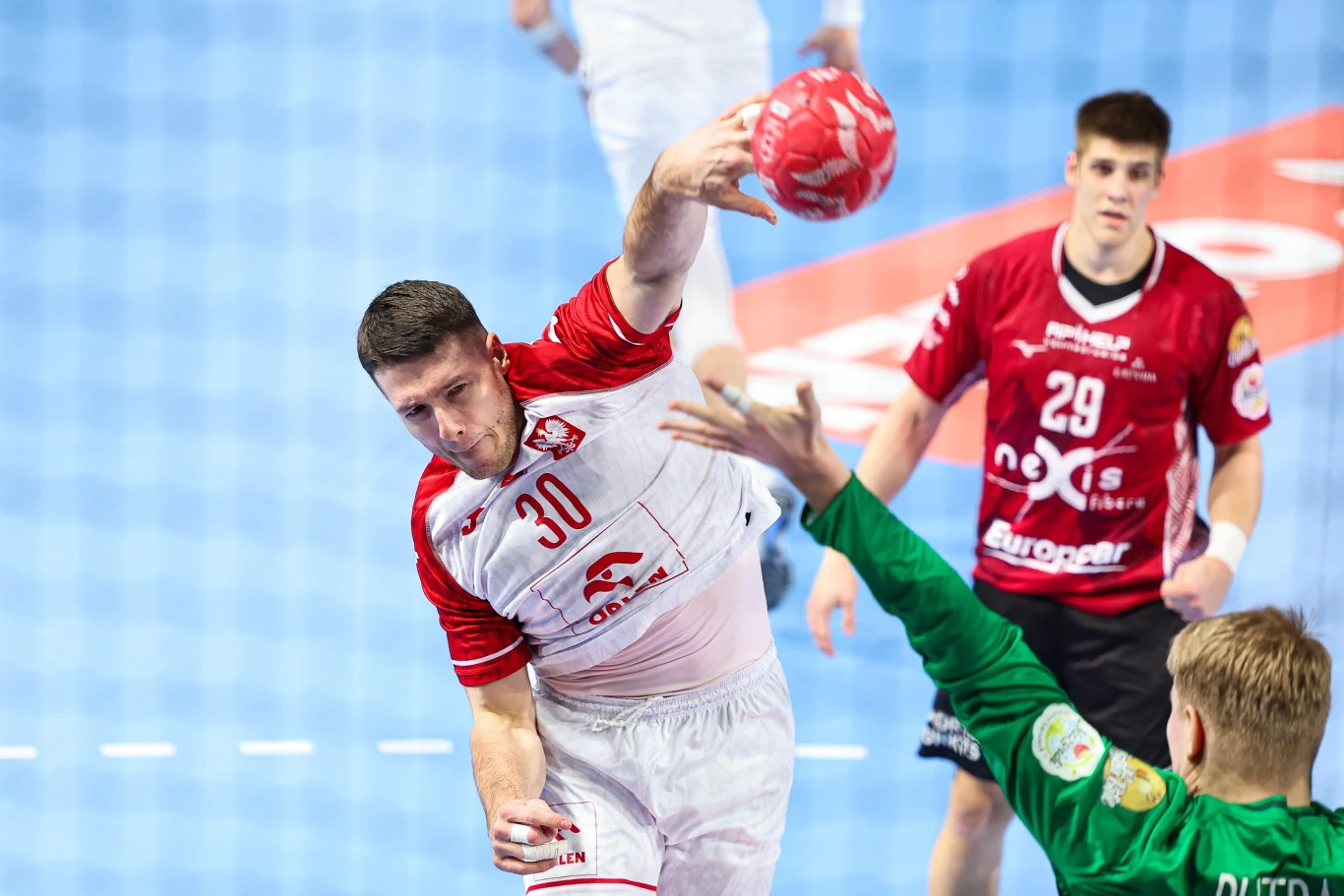 zawodnik w białym stroju z numerem 30 wykonuje rzut podczas meczu piłki ręcznej, atakując bramkarza drużyny w zielonym stroju; w tle widoczny jest zawodnik przeciwników w czerwonej koszulce z numerem 29