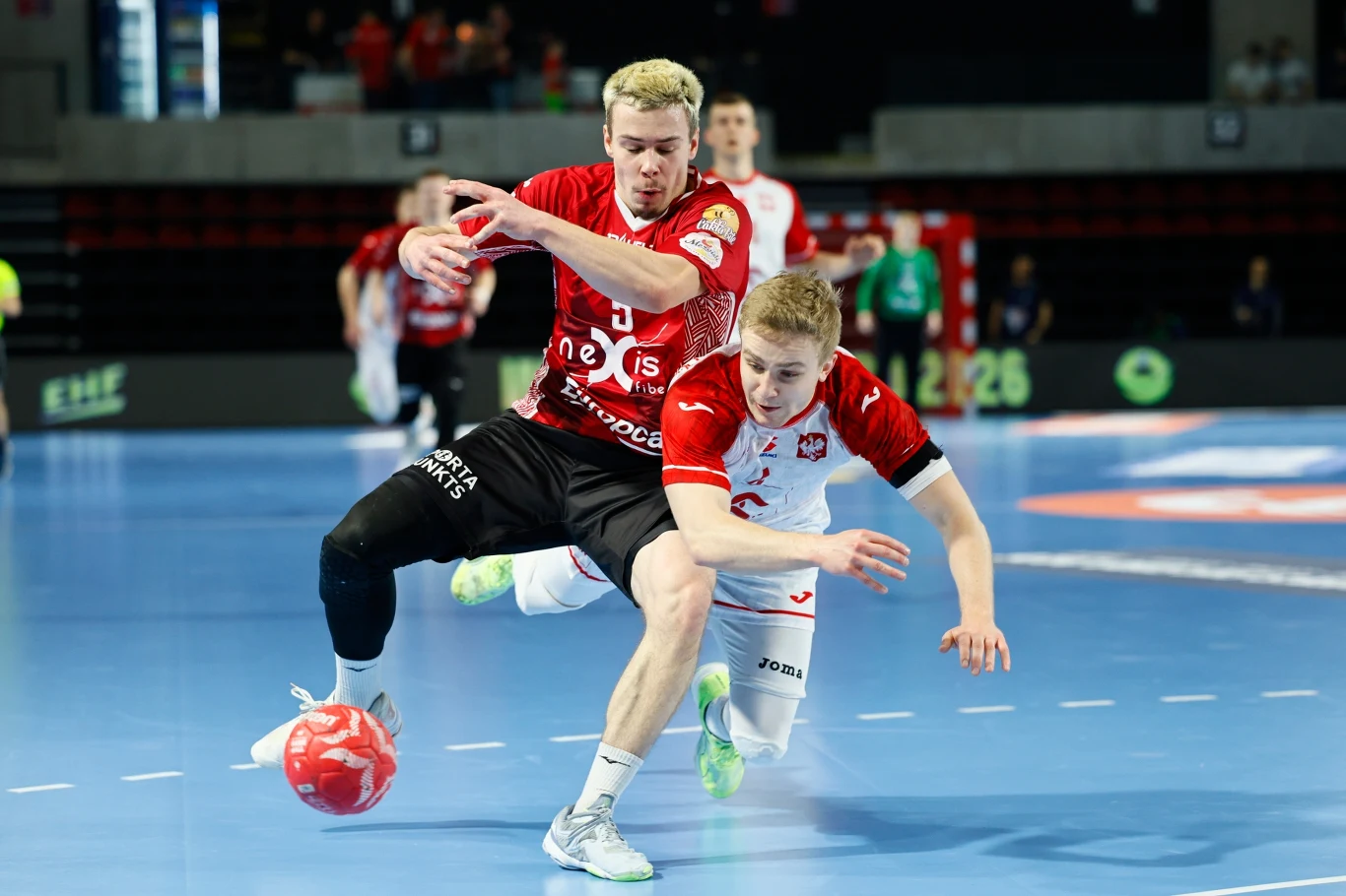 Dwóch szczypiornistów w dynamicznej walce o piłkę podczas meczu piłki ręcznej na hali, jeden zawodnik w czerwono-czarnej koszulce próbuje przejąć piłkę, podczas gdy drugi w biało-czerwonej koszulce upada na parkiet, w tle widać rozmazanych zawodników i...