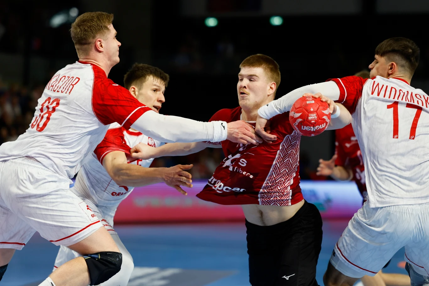 dynamiczna walka podczas meczu piłki ręcznej, zawodnik w czerwonej koszulce próbuje przedrzeć się przez obronę trzech zawodników w białych strojach, trzymając piłkę w prawej ręce