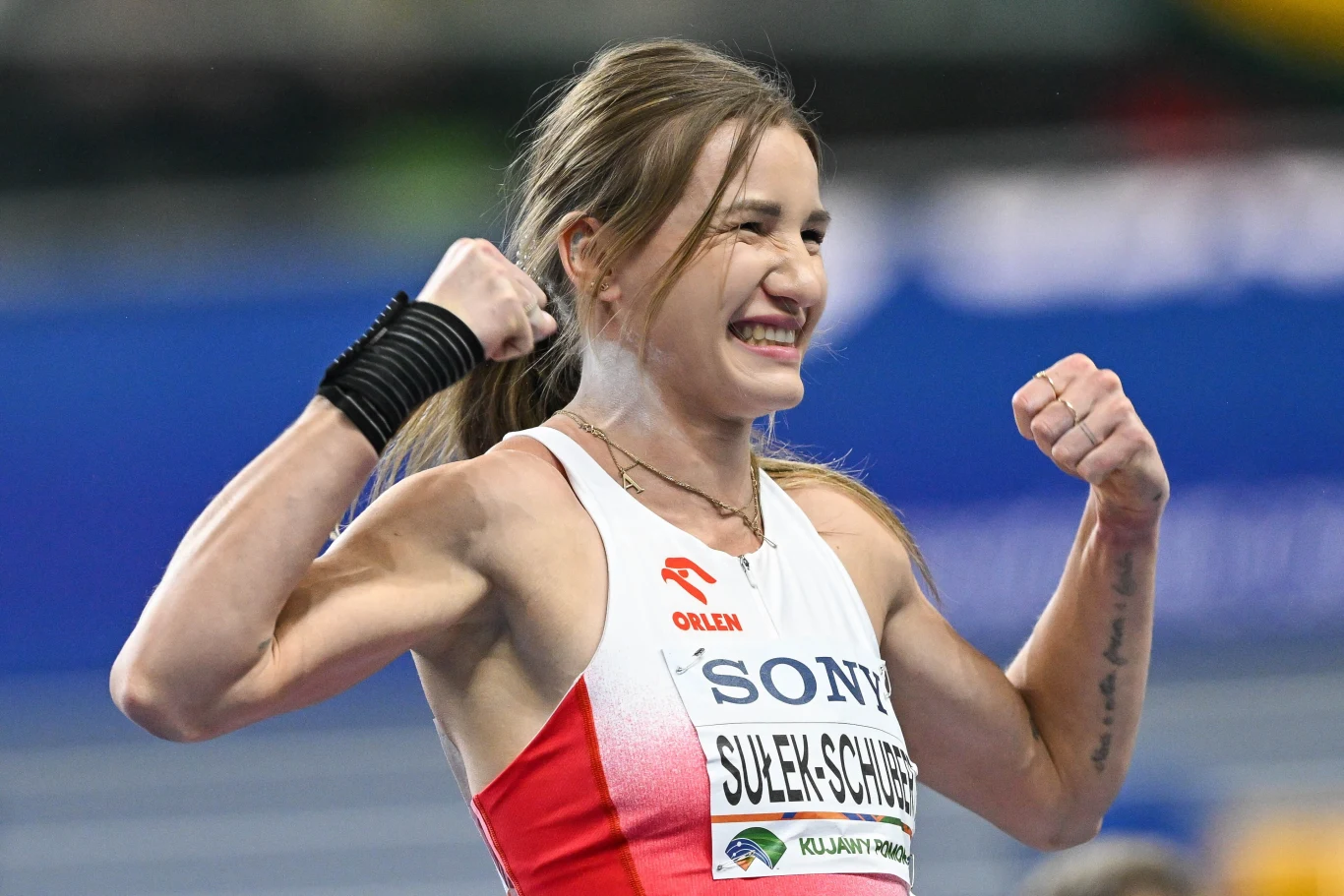 Lekkoatletka w biało-czerwonym stroju sportowym z nazwiskiem na koszulce świętuje sukces, unosząc zaciśnięte pięści i uśmiechając się szeroko, na tle niebieskiej bieżni stadionu.