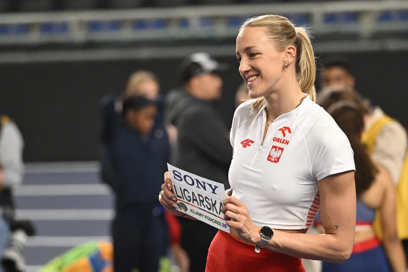 kobieta w stroju sportowym reprezentacji Polski, trzymająca tabliczkę startową z nazwiskiem i uśmiechnięta w halowej hali sportowej