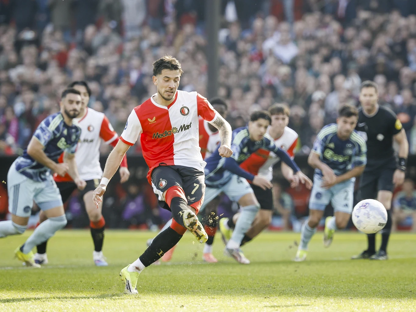 Piłkarz drużyny Feyenoord wykonuje rzut karny podczas meczu, skoncentrowany na piłce, podczas gdy zawodnicy obu drużyn obserwują sytuację i przygotowują się do ewentualnej interwencji.