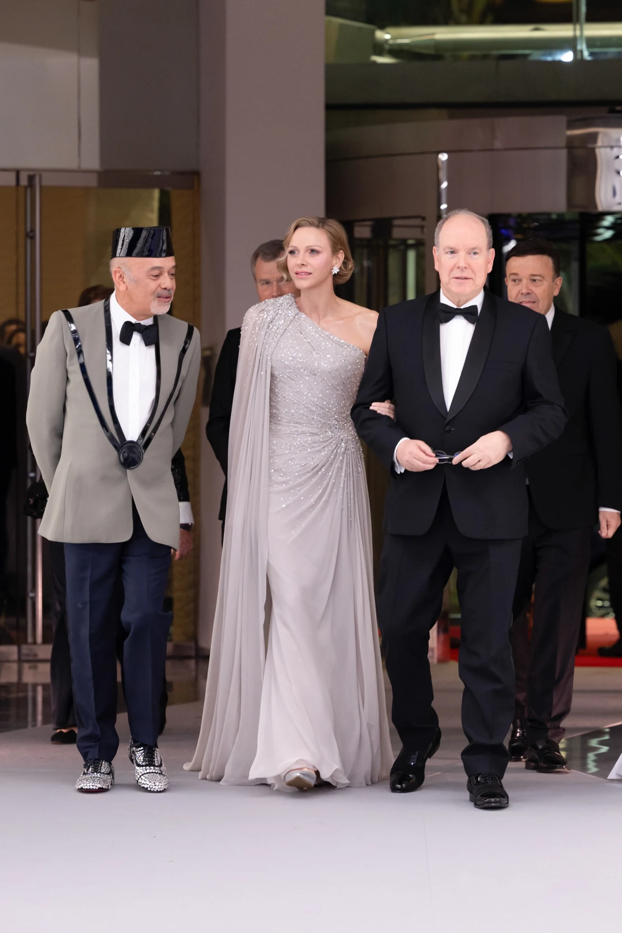 Elegancko ubrani mężczyźni w smokingu oraz kobieta w długiej, srebrzystej sukni idą razem po czerwonym dywanie, przyciągając uwagę otoczenia swoim szykiem i stylem.