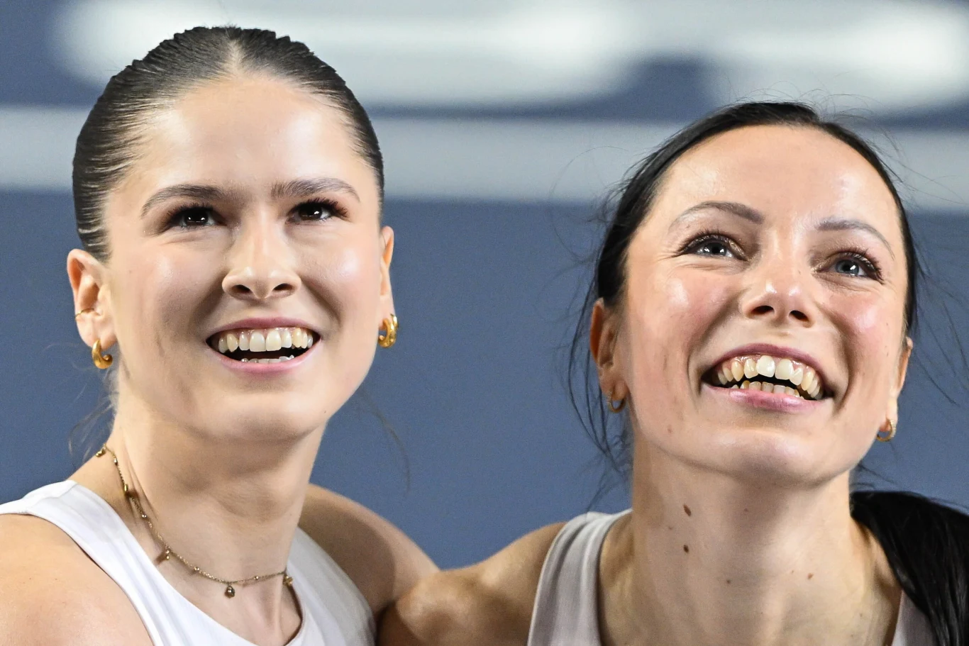 Dwie kobiety z szerokimi uśmiechami stoją obok siebie, ubrane w jasne sportowe topy, wyglądają szczęśliwie i radośnie w jasnym oświetleniu.