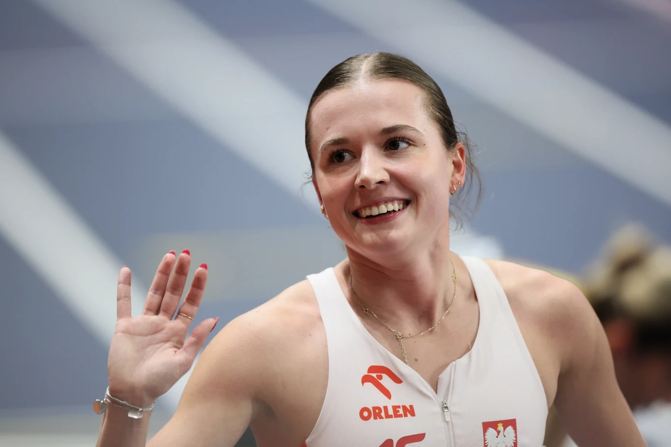 Uśmiechnięta sportsmenka w białym stroju z godłem Polski i logo sponsorów, z uniesioną dłonią w geście powitania lub gratulacji, na tle bieżni lekkoatletycznej.