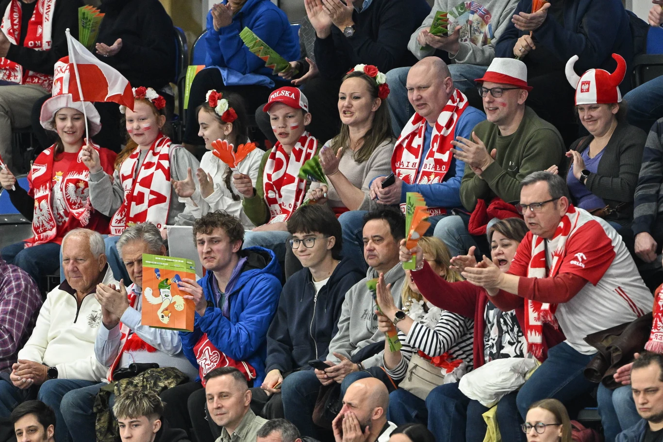 Grupa kibiców ubranych w barwy Polski, z szalikami, czapeczkami i akcesoriami w kolorach biało-czerwonych, dopingujących drużynę podczas wydarzenia sportowego na trybunach.