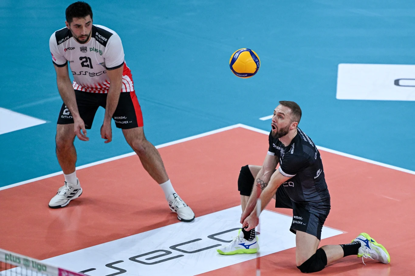 zawodnik w czarnej koszulce wykonuje odbiór piłki do siatkówki, klęcząc na kolorowym boisku, za nim stoi przeciwnik w biało-czerwonej koszulce, a żółto-niebieska piłka znajduje się tuż nad jego ramieniem