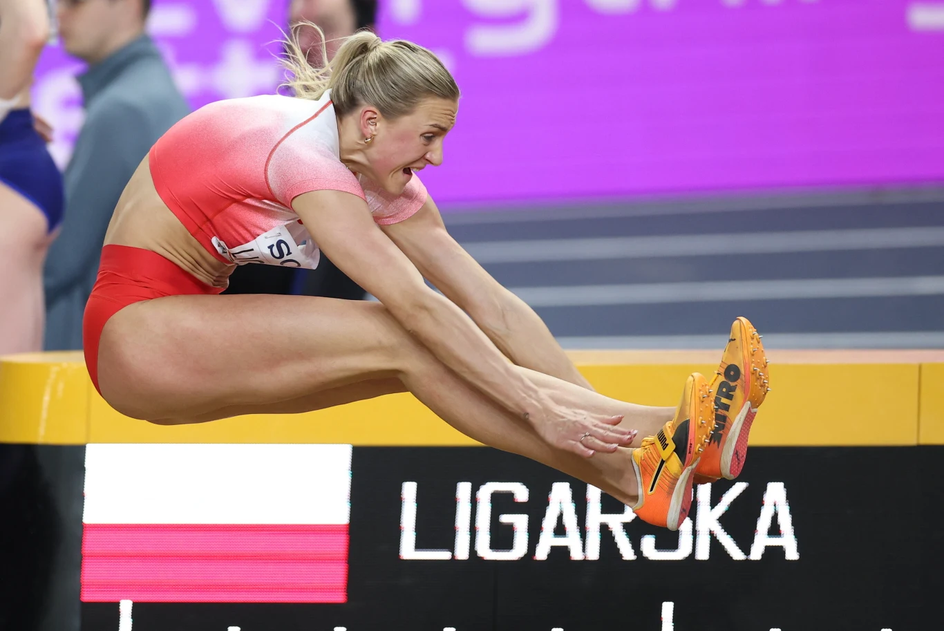 lekkoatletka w czerwono-białym stroju wykonuje skok w dal, unosząc ramiona oraz zgięte nogi, w tle tablica z napisem 'LIGARSKA' i polską flagą