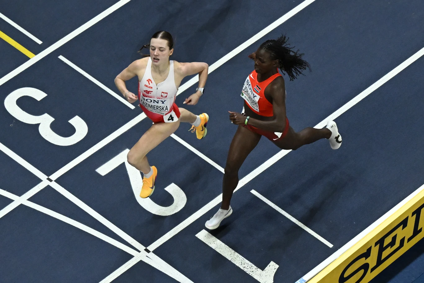 Dwie zawodniczki przekraczają linię mety podczas biegu na hali; pierwsza biegnie w biało-czerwonym stroju reprezentacji Polski, druga w pomarańczowym stroju reprezentacji Holandii, obie w wyraźnym wysiłku fizycznym, z dynamicznym ruchem ramion i nóg.