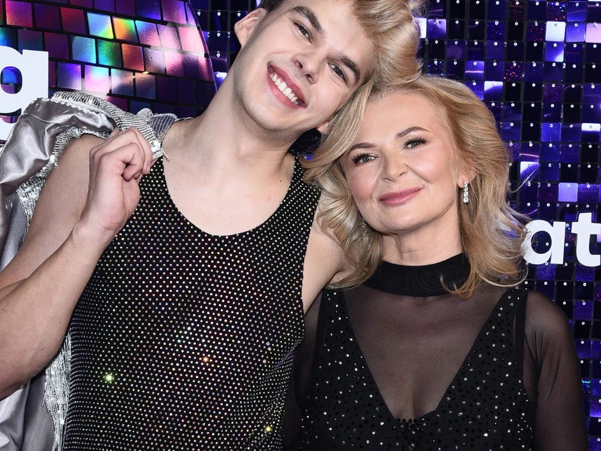 Uśmiechnięta para pozuje razem na tle połyskującej, fioletowej ściany z błyszczącymi kwadratami; obie osoby mają na sobie czarne, ozdobione cekinami stroje, a ich gesty i wyraz twarzy wyrażają bliskość i radość.