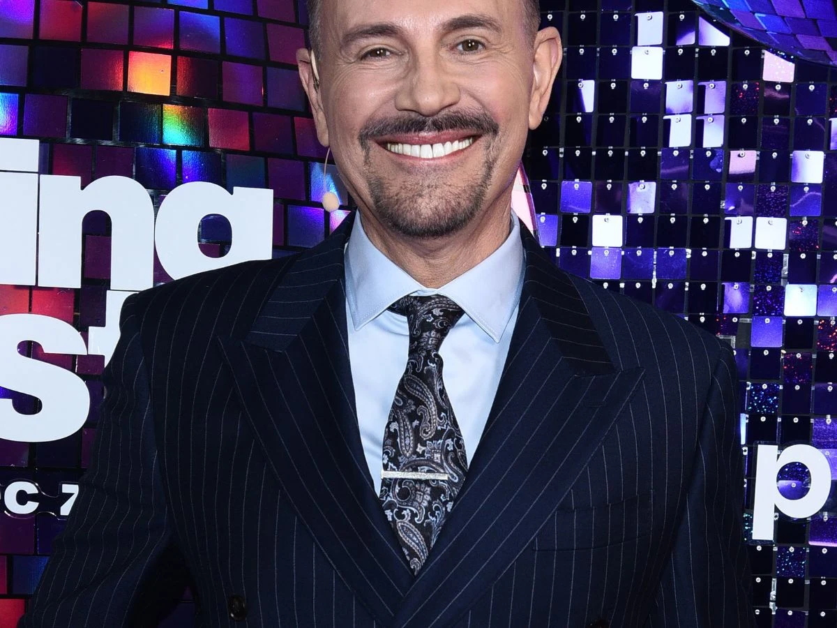 uśmiechnięty mężczyzna w eleganckim, granatowym garniturze w prążki, jasnoniebieskiej koszuli i ozdobnym krawacie na tle błyszczącej, kolorowej scenografii z elementami lśniących kafelków i fragmentami napisu