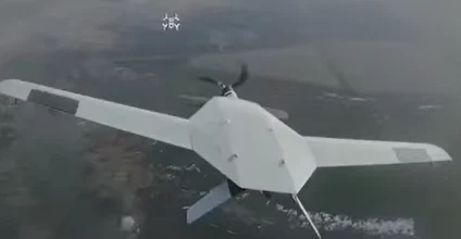 duży bezzałogowy dron o białym korpusie i prostokątnych skrzydłach leci nad rozległym, zamglonym terenem