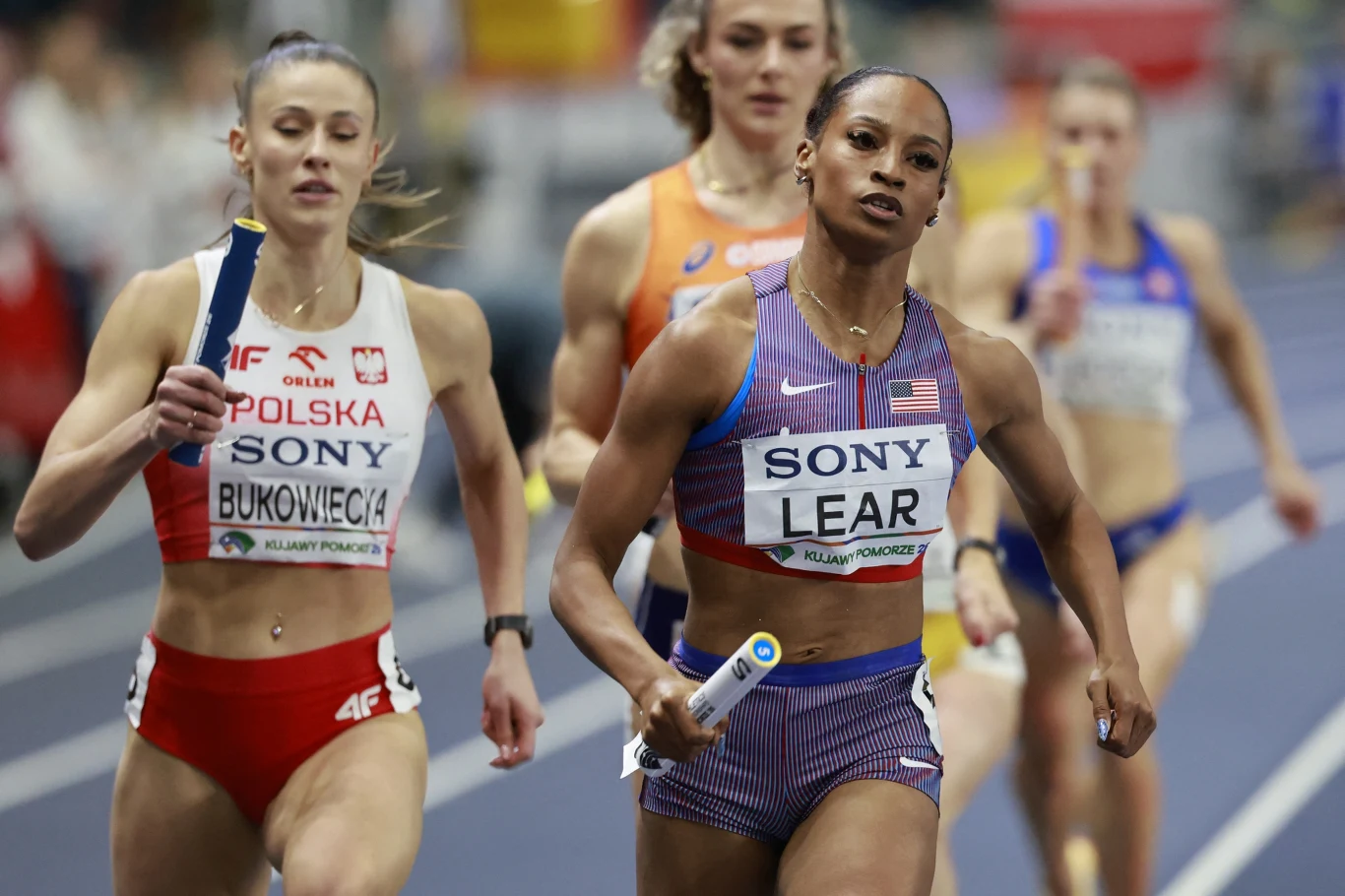 Zawodniczki z różnych krajów biegną na bieżni podczas sztafety, każda z nich trzyma pałeczkę sztafetową; na pierwszym planie reprezentantka USA i Polski rywalizują o prowadzenie.