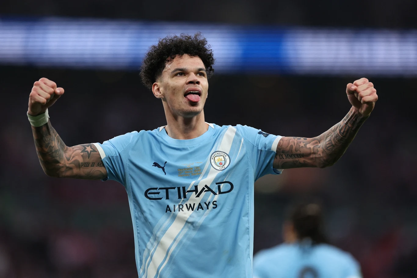 piłkarz Manchesteru City w jasnoniebieskiej koszulce z uniesionymi pięściami, pokazuje wyraz radości i satysfakcji, wystawia język w geście triumfu, w tle rozmazane sylwetki innych zawodników