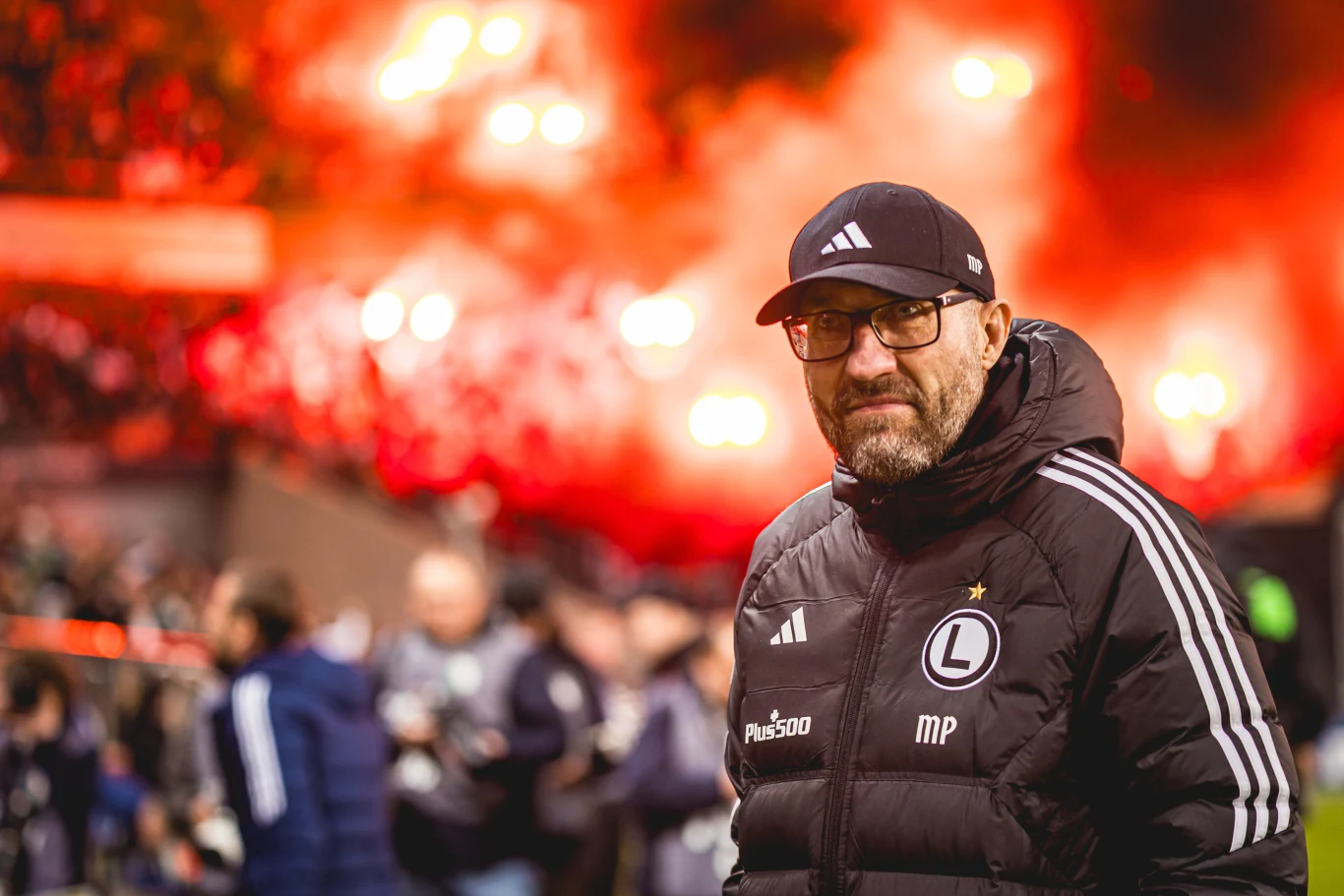 mężczyzna w sportowej kurtce i czapce z daszkiem, stojący na stadionie piłkarskim, na tle czerwonych rac dymnych i jasnych świateł na trybunach