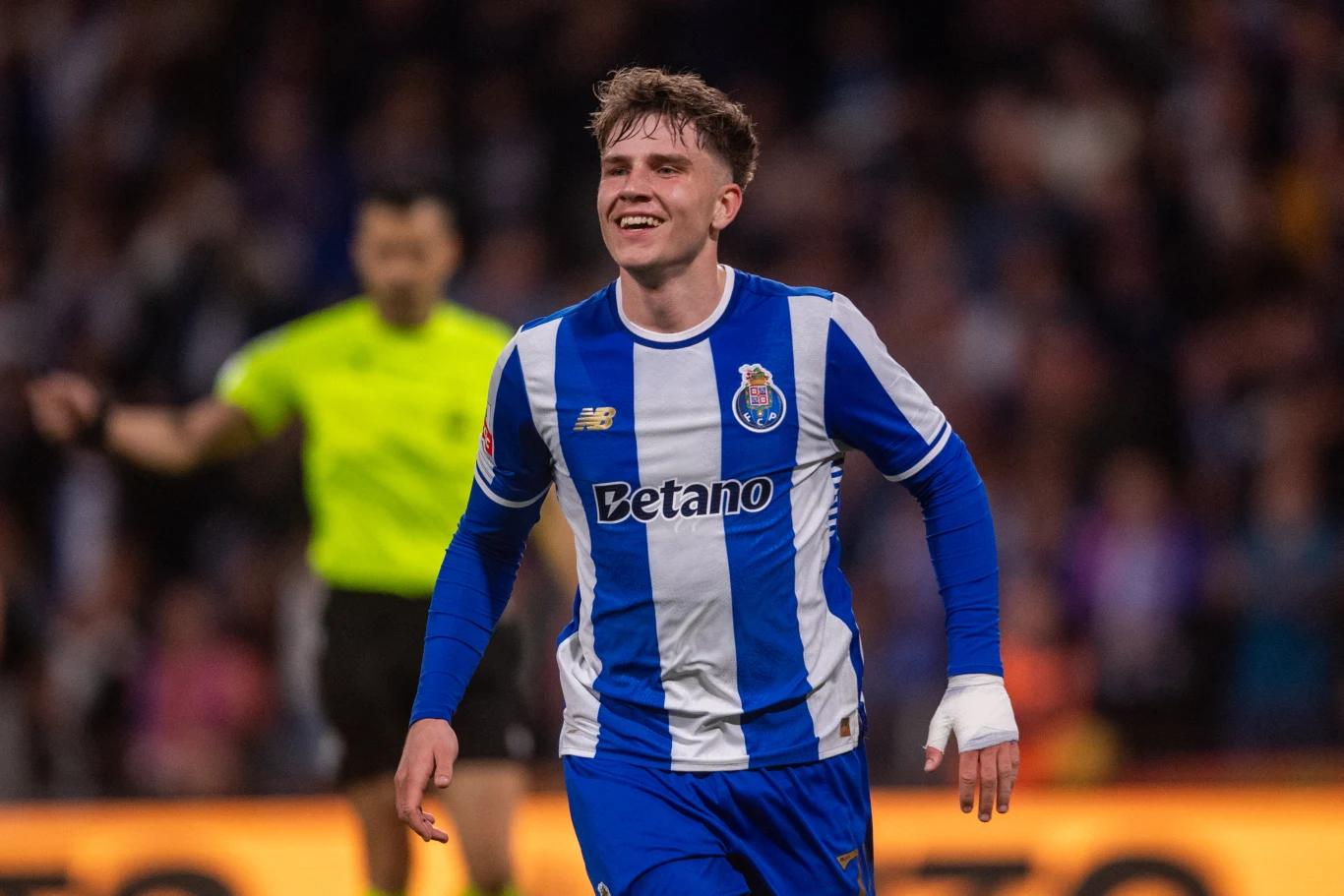 piłkarz w niebiesko-białej koszulce FC Porto biegnie po boisku z uśmiechem na twarzy, w tle widoczny sędzia w żółtym stroju