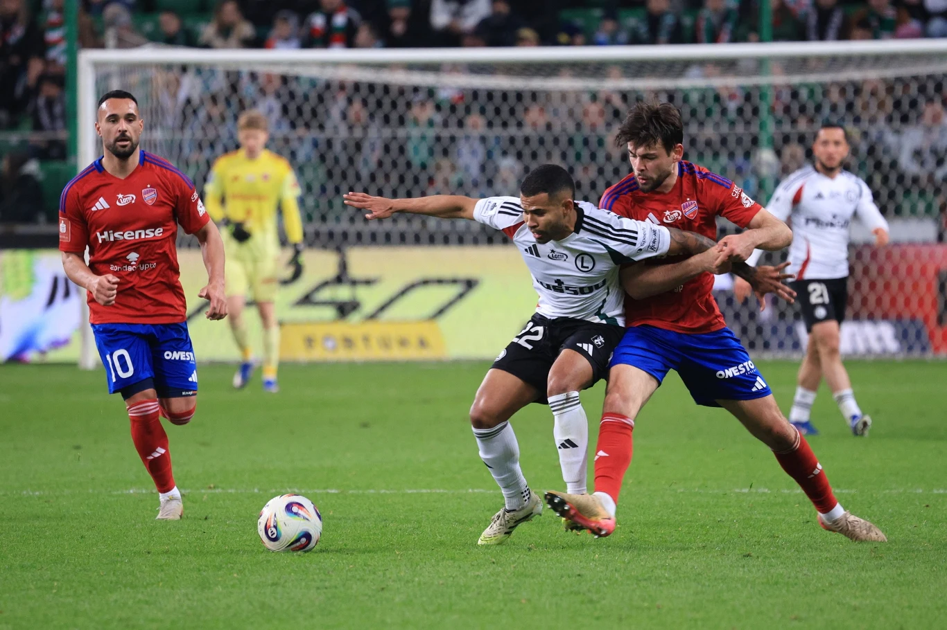 intensywna walka o piłkę pomiędzy zawodnikiem w białym stroju a piłkarzem w czerwonej koszulce, skoncentrowana w centrum akcji, podczas meczu piłkarskiego na zielonej murawie; w tle widoczni pozostali zawodnicy oraz bramkarz