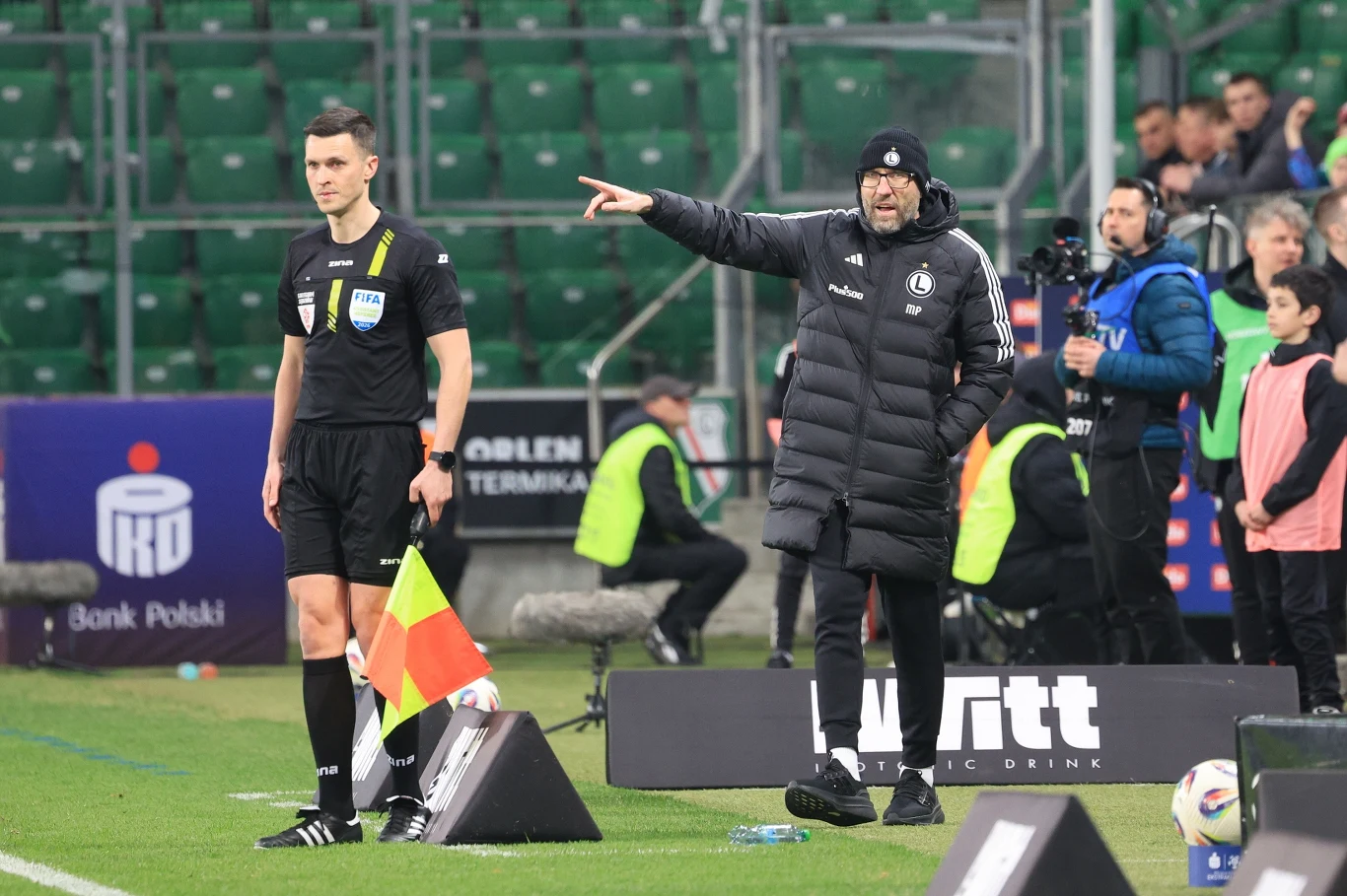 trener w czarnej kurtce i czapce wydaje polecenia z linii bocznej boiska, obok stoi sędzia boczny z chorągiewką, w tle widoczne są zielone trybuny i kilku pracowników technicznych